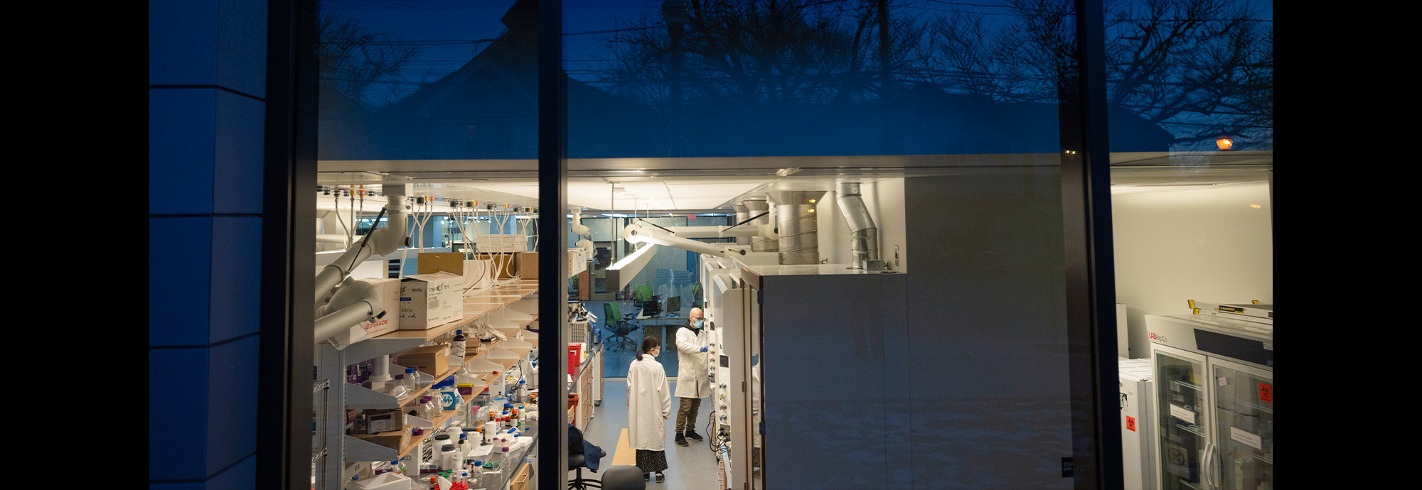 External of building looking through a window to two people working in a lab
